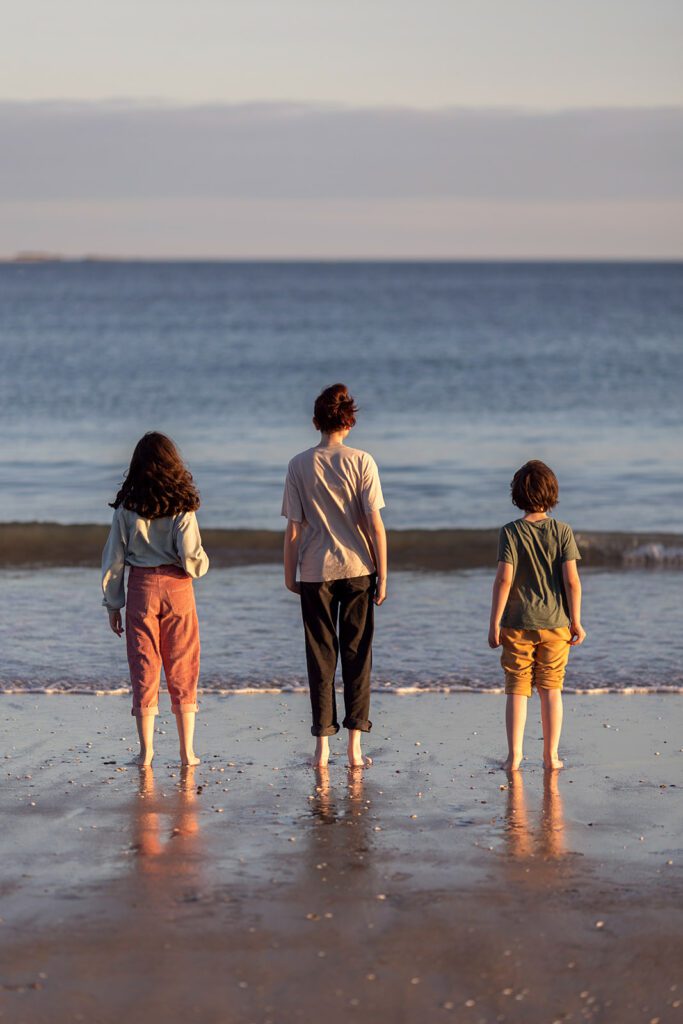 three siblings looking at the sea waves, photographed from the back by a documentary family photographer