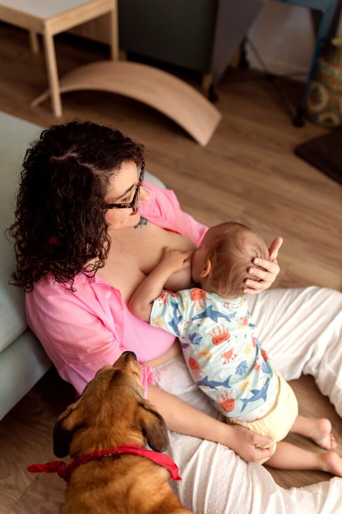 a mom is breastfeeding her toddler at home with her dog sitting next to them, photographed by a documentary family photographer