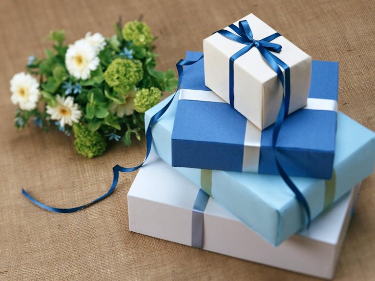 Three beautifully wrapped gift boxes with blue ribbons arranged beside a floral bouquet, Gift ideas for parents of a newborn