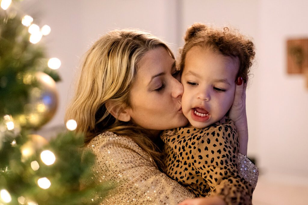 mom kissing her little girl near the family christmas tree during a christmas family photo session at home