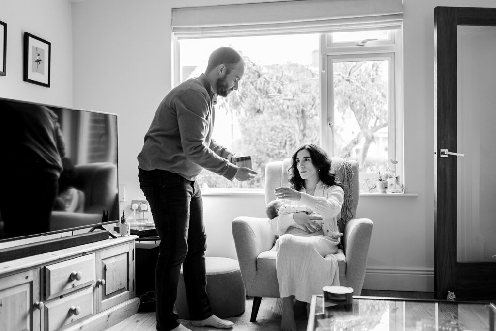 father giving his wife a cup of thea while she nurses their newborn at home