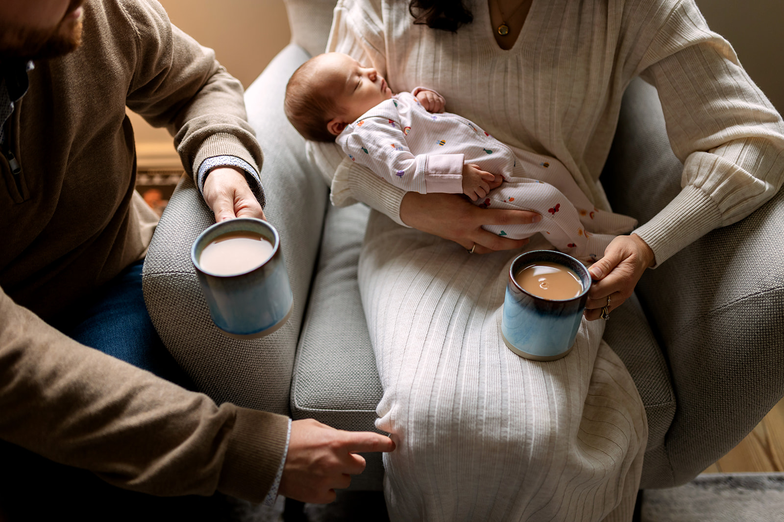 Parents sitting together with cups of tea and their newborn sleeping in mum's arms